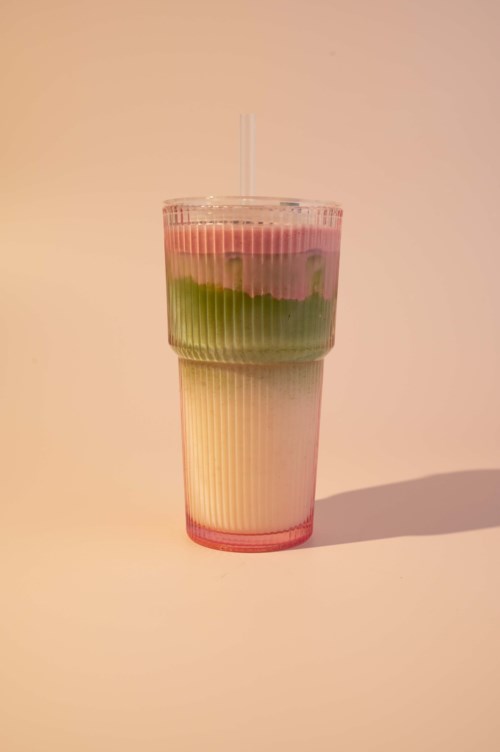 En lagd drink med rosa, grønt og hvitt i et klart plastglass med sugerør, på en enkel bakgrunn.