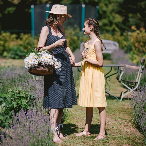Een vrouw en een meisje praten in een tuin met paarse bloemen, vrouw houdt een mand met madeliefjes vast, zonnige dag, casual kleding.