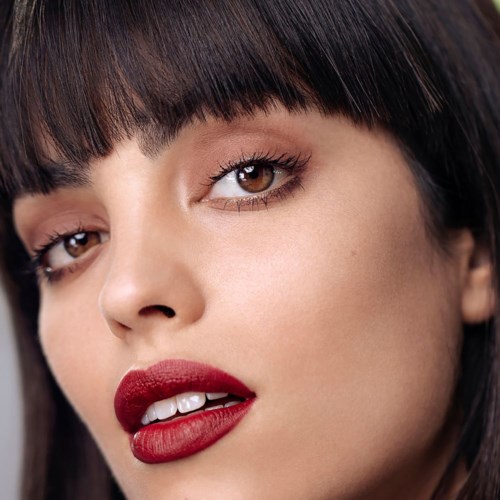 Close-up van een vrouw met bruine ogen, donker haar en rode lippenstift die haar tanden licht laten zien.