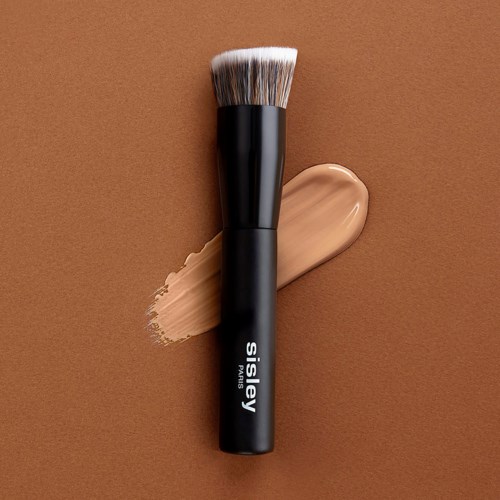 Een make-upkwast met een zwarte handvat ligt op een bruine achtergrond, met beige foundation eronder.