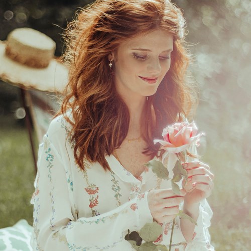 A woman with red hair gently holds a pink rose, standing outdoors in sunlight, wearing a light-colored floral dress.
