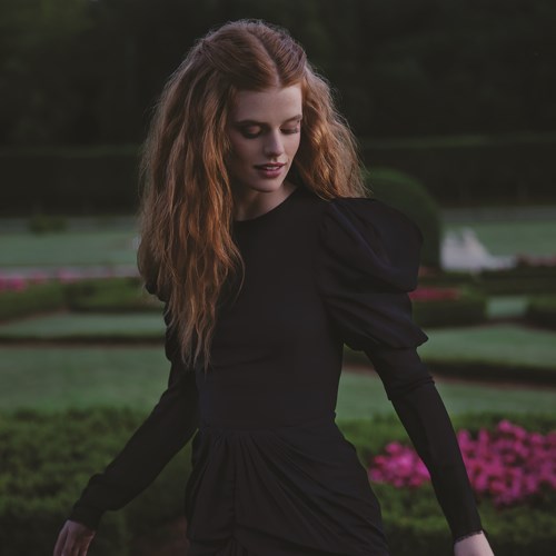 Een jonge vrouw met lang, krullend haar staat in een tuin met roze bloemen en groen gras.