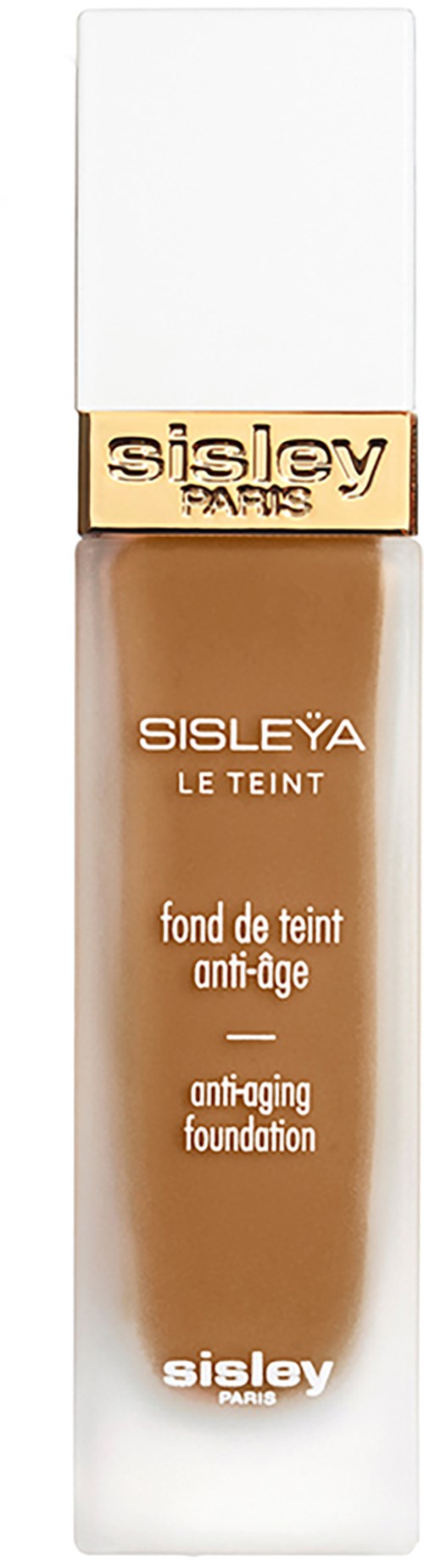 A tall, slim bottle with a white cap, gold band, and beige foundation inside. Text on bottle includes brand and product details.