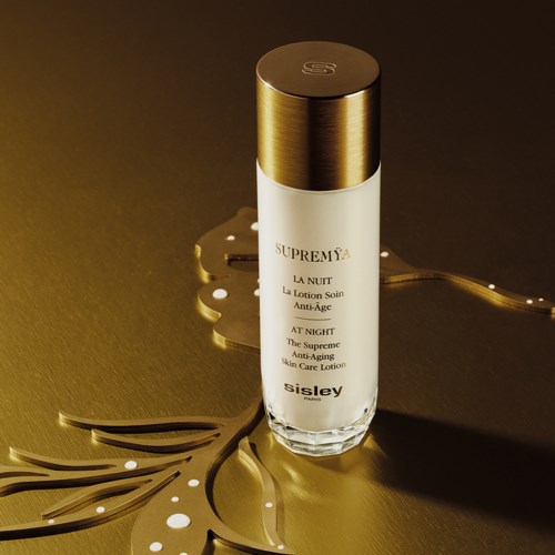 Eine weiße zylindrische Flasche mit goldener Kappe und goldenen Schriftzügen, auf dunklem Hintergrund mit einem dekorativen goldenen Element.