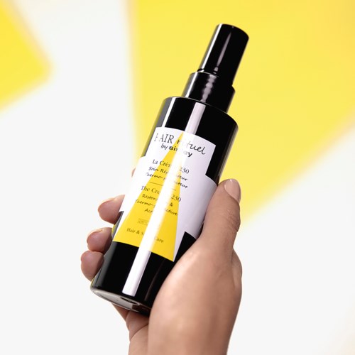Een hand houdt een zwarte sprayfles met een wit label en gele accenten, tegen een heldergele achtergrond.