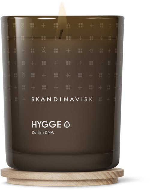 Dunkle Kerze in Glas mit skandinavischen Mustern, beschriftet 'HYGGE' und 'Danish DNA,' auf einem Holzuntersetzer.