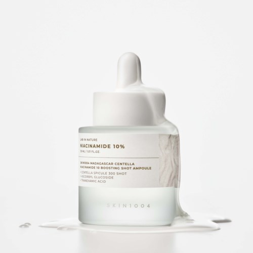 A clear glass bottle with a white dropper cap, containing a skincare serum labeled Niacinamide 10%. Some serum is spilled around.