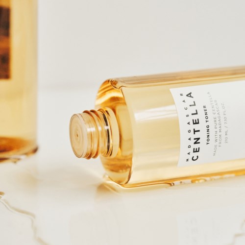 Eine klare Flasche mit Gesichtswasser und weißem Etikett, liegt auf einer weißen Oberfläche. Eine andere Flasche ist teilweise im Hintergrund sichtbar.