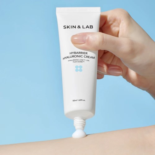Een hand drukt witte crème uit een tube met 'Skin & Lab' op een blauwe achtergrond.