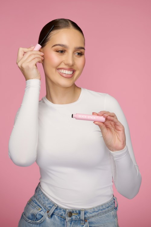 Een lachende vrouw in een wit shirt houdt roze ontharingscrème. Ze gebruikt een spatel en heeft een roze achtergrond.