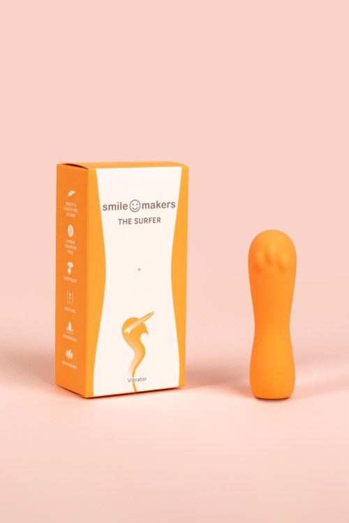 Ein hellorangener, formschöner Gegenstand neben einem Karton mit der Aufschrift "Smile Makers The Surfer" auf rosa Hintergrund.