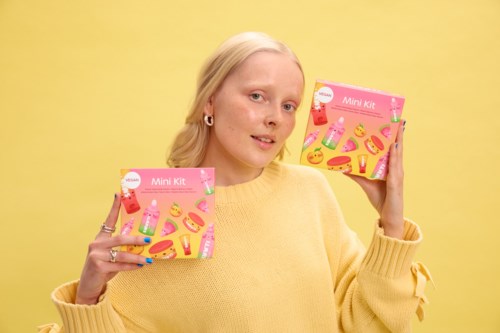 A young woman with blonde hair in a yellow sweater holds two pink mini kit boxes, standing in front of a yellow background.