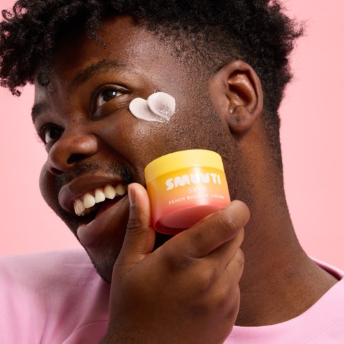 Een man met krullend haar die lacht, houdt een potje met crème, met crème op zijn wang, tegen een roze achtergrond.