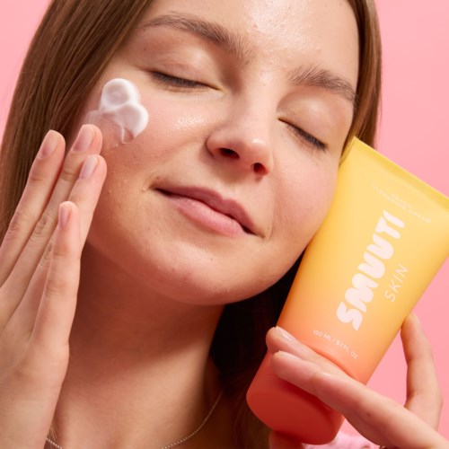 Eine Frau trägt Creme ins Gesicht auf, hält ein gelbes Hautpflegeprodukt vor rosa Hintergrund.
