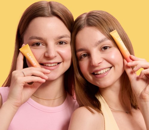 Twee meisjes met lichtbruin haar die gele huidverzorgingsproducten vasthouden en glimlachen tegen een gele achtergrond.