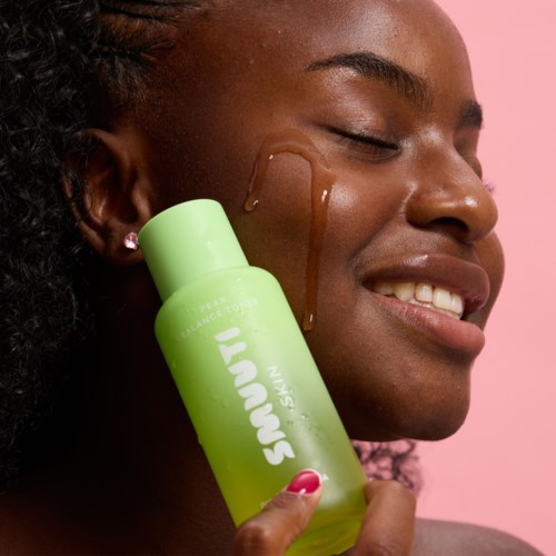 Een lachende vrouw heeft een druppel huidverzorgingsproduct op haar wang en houdt een groene fles met 'Sun Bum Skin'.