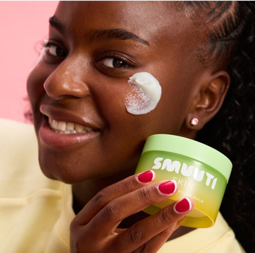 Eine Frau lächelt und trägt Creme auf die Wange auf, während sie einen grünen Behälter mit Smuti Skin hält.