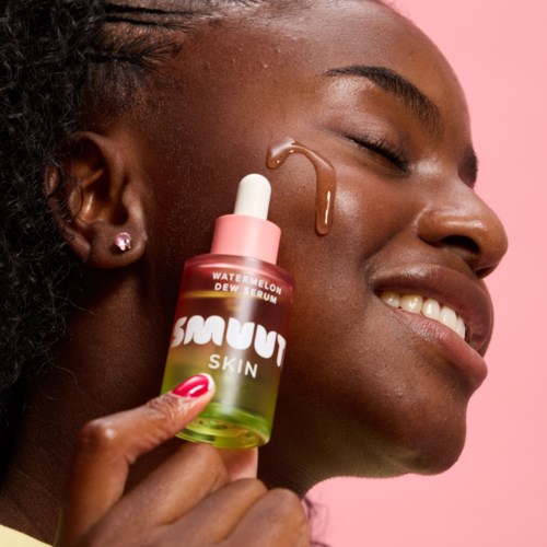 Een vrouw glimlacht met een druppel huidserum op haar gezicht en houdt een klein flesje met "Watermelon Dew Serum."