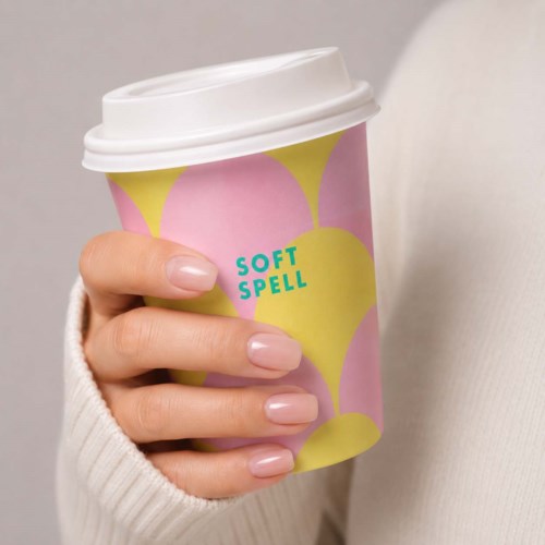 En hand håller en rosa och gul papperskopp med vit lock, märkt Soft Spell.