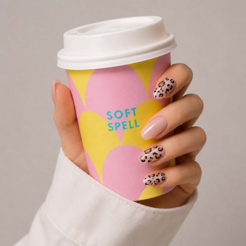 En hand med leopardmönstrade naglar håller en färgglad pappersmugg med vit lock, märkt Soft Spell.