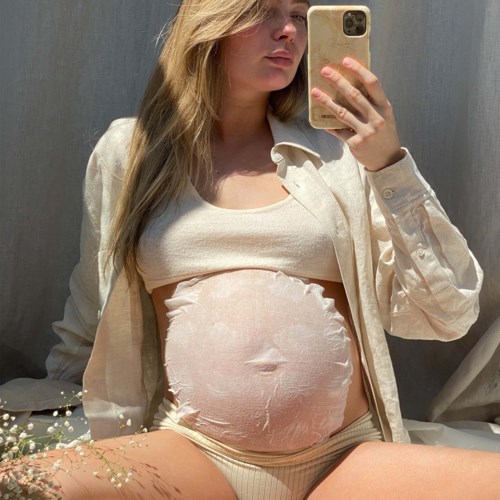 Eine schwangere Frau macht ein Selfie im Spiegel. Sie trägt ein helles Hemd offen über ihrem Babybauch und ein passendes Top.