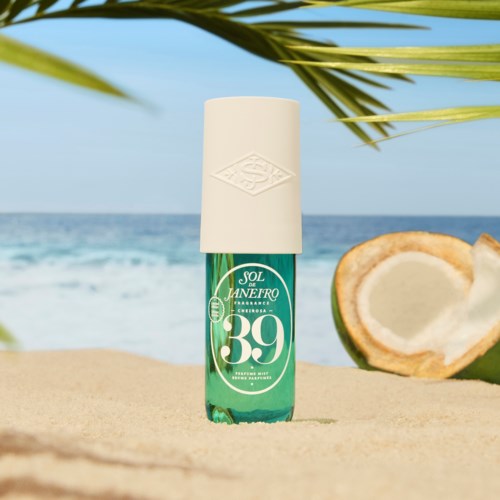 A bottle of spray sunscreen on a beach with a coconut and ocean in the background.