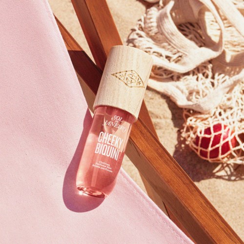 Eine rosa Flasche mit der Aufschrift Cheeky Biquin, Sonnenlicht, Holzmöbel, Stoff und eine Netztasche sind sichtbar.
