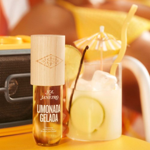 Kleine Flasche mit dem Aufdruck Limonada Gelada neben einem Glas mit Zitronenscheiben und Strohhalm auf gelber Oberfläche.