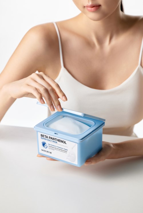 En kvinna i vit topp håller en blå behållare märkta 'Beta Panthenol' på en vit yta.