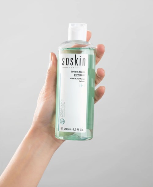 A hand holds a clear bottle of Soskin gentle purifying toner against a plain background.