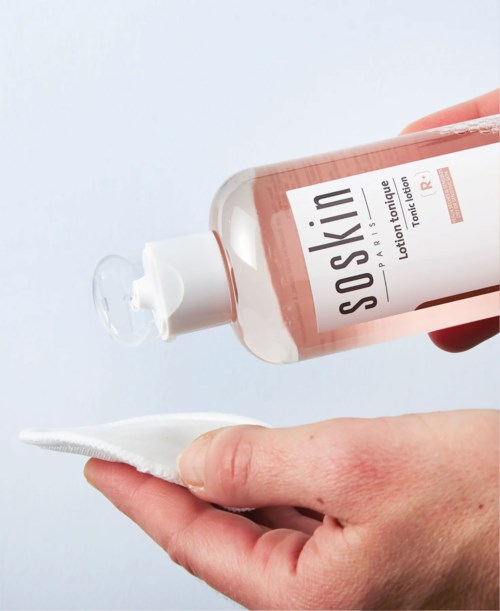A hand holds a bottle of SoSkin toner and a cotton pad, ready to apply the product.