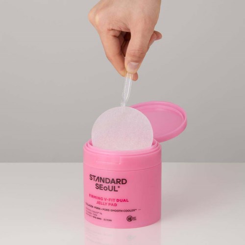 A hand lifts a white cleansing pad from a pink container labeled 'STANDARD SEOUL.'
