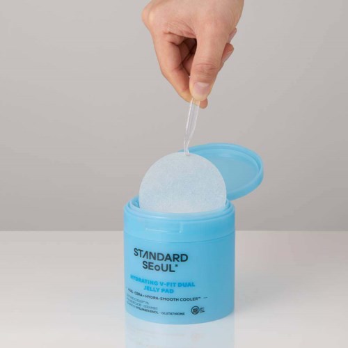 A hand dips a dropper into a blue container labeled 'Standard Seoul' skincare product, with a cotton pad inside.