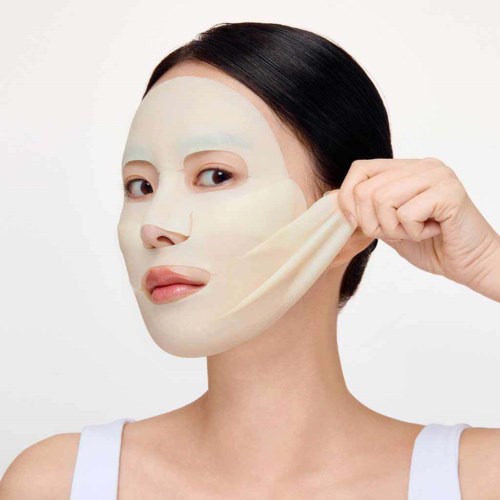 A woman peels off a facial mask sheet, revealing smooth skin beneath, with a neutral facial expression.