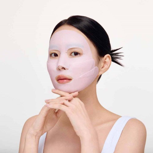 A woman applies a white facial mask, focusing on her face, with dark hair tied back and a neutral background.
