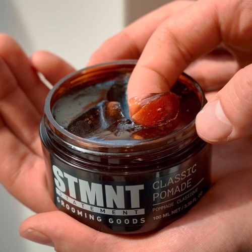 Hands dip fingers into a jar of grooming pomade, labeled 'STMT Classic Pomade.' The pomade is dark and thick.