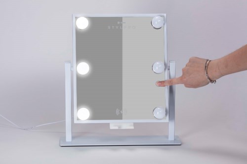 Een spiegel met lichten eromheen, een hand raakt een licht aan, en er is een standaard en netsnoer.