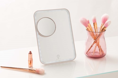 Een witte make-up spiegel met een ronde vergrotende spiegel, naast roze make-up borstels in een roze vaas.