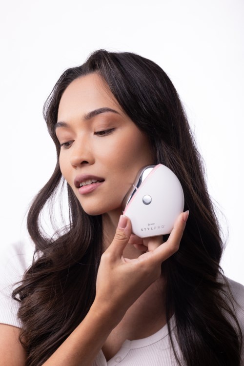 A woman with long dark hair uses a small white device against her cheek.