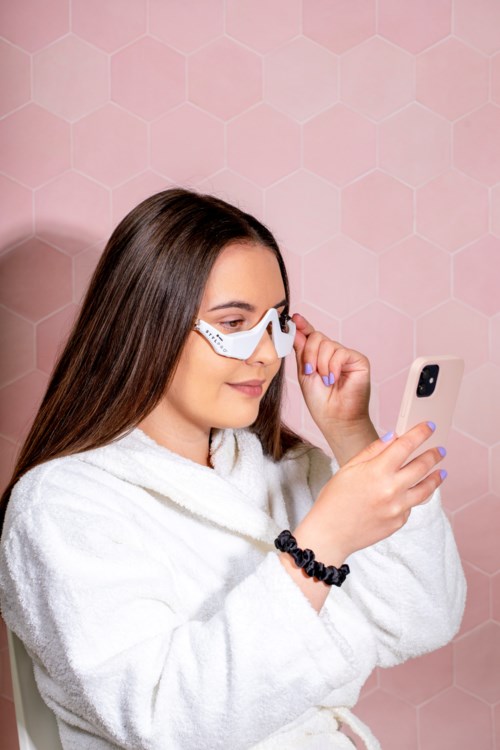 Een vrouw in een badjas kijkt naar haar telefoon tegen een roze honingraatachtergrond.
