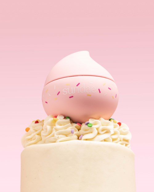 Ein Kuchen mit weißem Zuckerguss, verziert mit bunten Streuseln, gekrönt von einem pinken und weißen Swirl-Spielzeug, vor rosa Hintergrund.