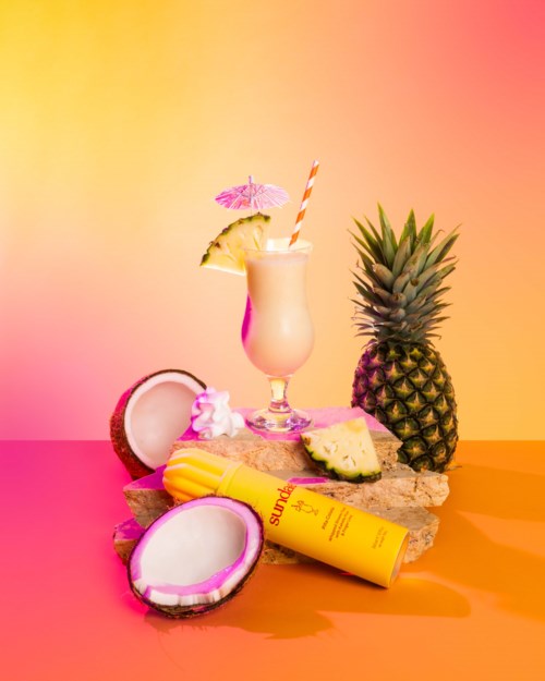 Bunte tropische Szene mit Ananas, Kokosnuss, Getränk und Hautpflegeprodukten vor pinkem und orangefarbenem Hintergrund.