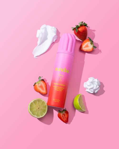 Roze zonnebrand spray omringd door aardbeien, limoenhelften en witte slagroom op een roze achtergrond.