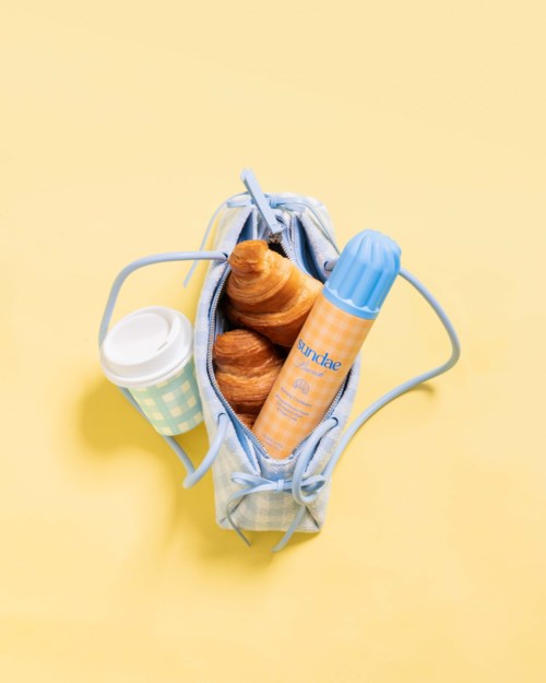 Een kleine tas met croissants, een drankje en zonnebrandcrème, op een gele achtergrond.