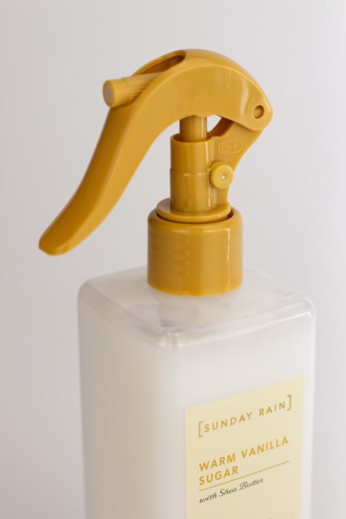 En vit sprayflaska med gul topp och utlösare, märkt Sunday Rain Warm Vanilla Sugar with Shea Butter.
