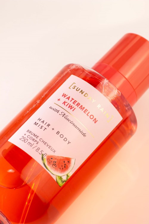 Ljus orange flaska av SunDew vattenmelon och kiwi hårmist med makadamia, ligger på en ljus yta.