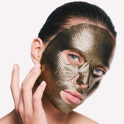 En kvinne med blendende blå øyne har på seg en gullfarget maske med bikakemønster som dekker ansiktet hennes, og hun berører kinnet med hånden.