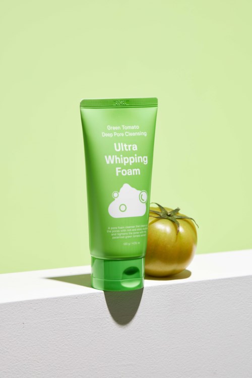 Groene tube met reinigingsschuim naast een gele tomaat op een wit oppervlak, achtergrond is lichtgroen.