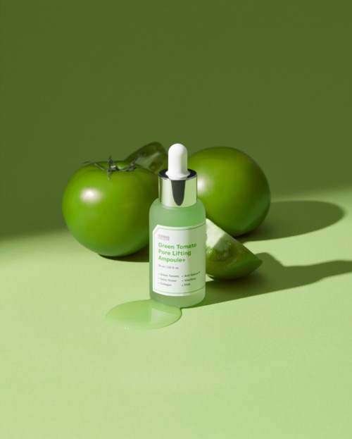 Een groen huidverzorgingsfles met druppelaar naast twee groene tomaten en een limoenwiel op een groene achtergrond.