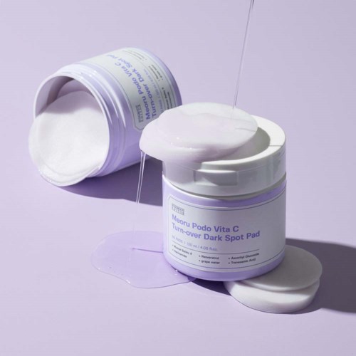 Een pot met label "Manu Podo Vita C Turn-over Dark Spot Pad" die room morst op een paarse achtergrond.